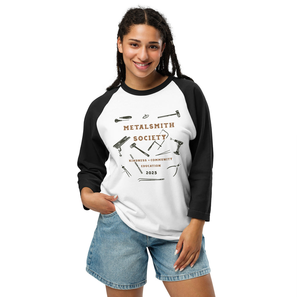 Smiling person wearing the Metalsmith Society's 2025 Raglan Shirt, featuring a white body with black raglan sleeves. The shirt showcases a graphic design of jewelry-making tools in black and metallic tones, along with the text 'Metalsmith Society - Kindness, Community, Education - 2025.' Paired with casual denim shorts, the shirt reflects a comfortable and creative style.