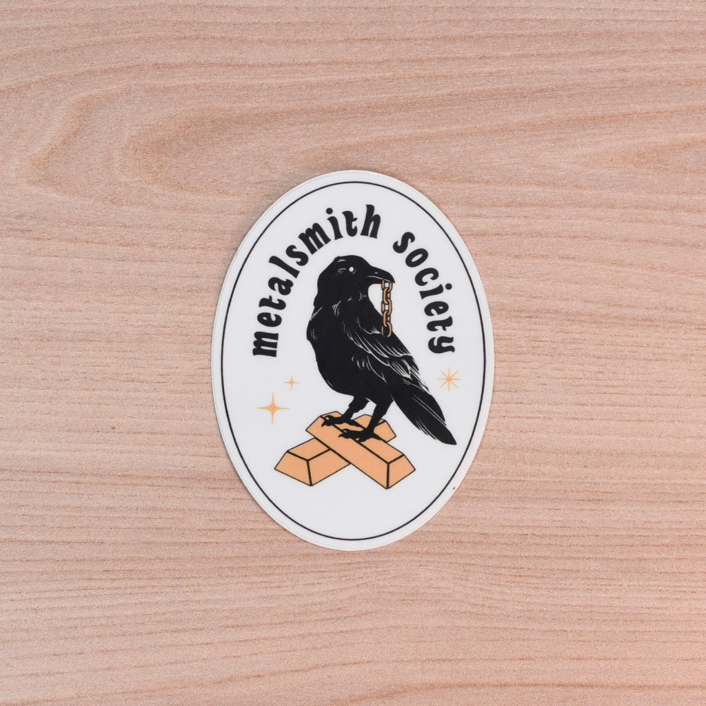 Sticker with a crow and ' Metalsmith society' text on a wooden surface