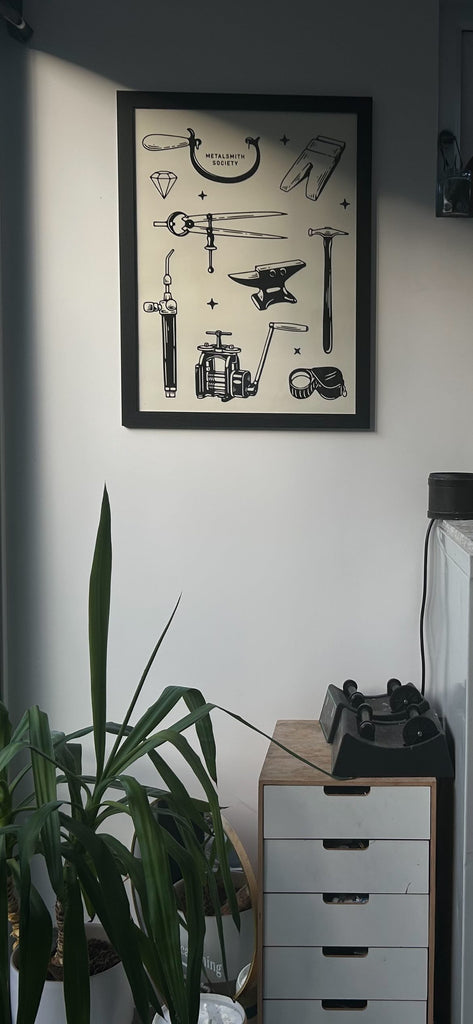 An image of the studio of @atypical.thing with a black framed print of jewelry making tools hanging on the wall and a plant in the corner.