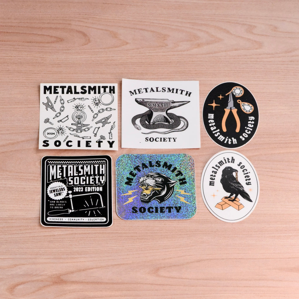 Metalsmith Society Jewelry Tool Sticker Pack featuring six unique designs from 2020–2024, including vinyl and glitter stickers displayed on a wooden surface.