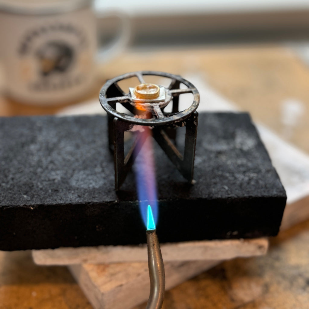  Ti Quad Soldering Stand in use, holding a gold bezel with a silver backplate and a torch flame can be seen heating from below. 