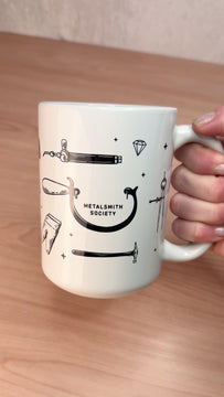 15oz Metalsmith Society ceramic mug featuring illustrated jewelry-making tools, tilting to show the design on a wooden surface.