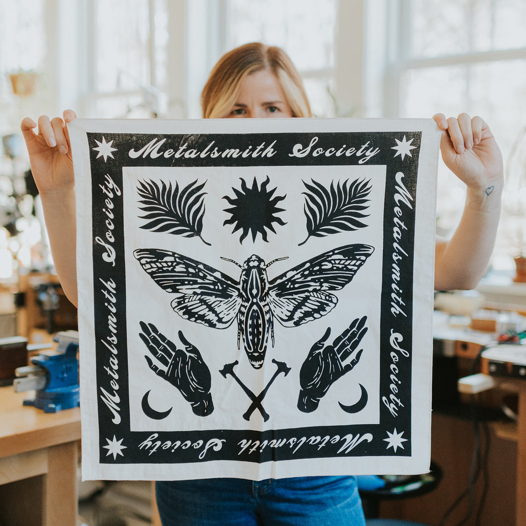 Close-up of the 2022 Bandana’s black ink pattern on natural fabric, highlighting jewelry tools and floral motifs. Being held in someone's hands.