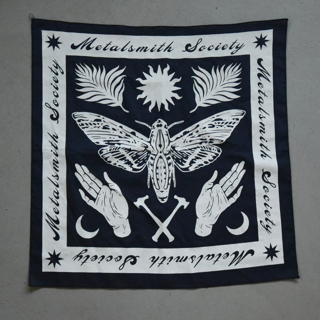 2022 Bandana laid flat, showcasing a navy fabric with detailed white ink illustrations of jewelry tools, floral, hands and moth designs.